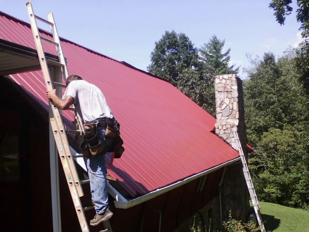 Roofer on a ladder beside a red metal roof and stone chimney — Soffit & Fascia Repair by Roofing Brothers in Idaho Falls